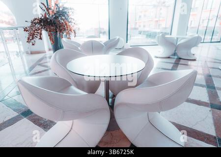 Moderner Loungebereich mit weißen skulpturalen Stühlen und rundem schwarzen Tisch auf Marmorboden, hell, geräumig und mit großem Glasfenster Stockfoto