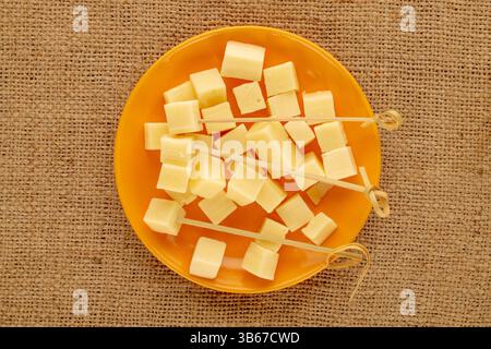 Duftender Hartkäse in Würfel geschnitten mit Bambusspießen auf einer Keramikplatte auf einem Jutetuch, Makro, Draufsicht. Stockfoto