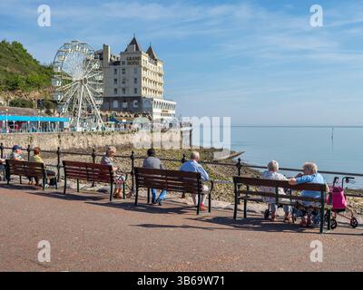 Senioren sitzen auf Promenadenbänken neben dem Pier in Llandudno, Nordwales, Großbritannien, in der warmen Frühlingssonne. Stockfoto