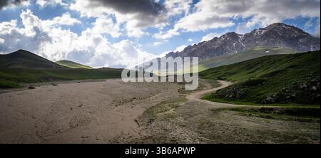 Eine malerische Landschaft mit sanften grünen Hügeln, einem sandigen Pfad und majestätischen Bergen im Hintergrund unter einem teilweise bewölkten Himmel. Stockfoto
