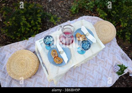 Gemütliches Picknick im Freien mit Gebäck, Beerensaft und blauen Gläsern auf einer Vintage-Tischdecke. Elegantes und rustikales Sommerrestaurant im Garten Stockfoto