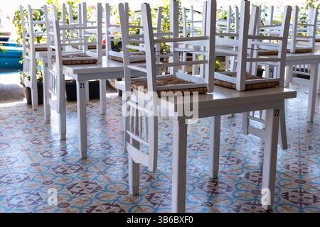 Weiße Holzstühle stapeln sich auf Tischen in einem geschlossenen griechischen Inselrestaurant und heben den farbenfrohen Fliesenboden hervor. Stockfoto