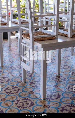 Weiße Holzstühle stapeln sich auf Tischen in einem geschlossenen griechischen Inselrestaurant und heben den farbenfrohen Fliesenboden hervor. Stockfoto
