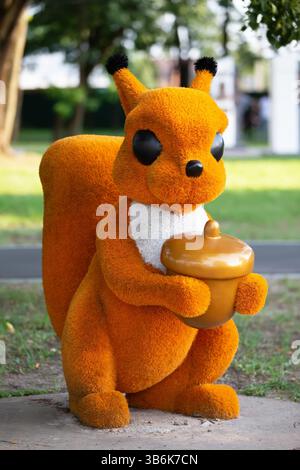 Ein großes orangenes Eichhörnchen hält eine falsche Eichel im Mund. Das Eichhörnchen ist aus unbekanntem Gras und steht auf einem grasbewachsenen Gebiet. Kleine architektonische Formen. Stockfoto