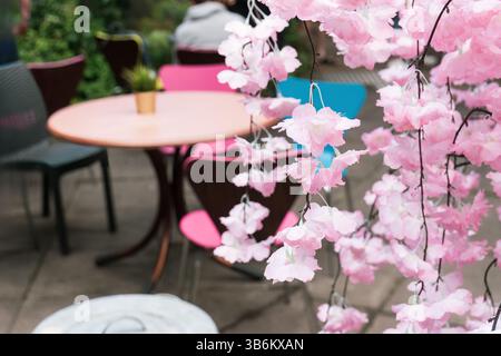 Rosafarbene künstliche Kirschblüten hängen im Vordergrund mit farbenfrohen Cafétischen im Freien im Hintergrund unscharf Stockfoto