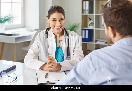 Männlicher Patient besucht eine freundlich lächelnde junge Ärztin, die am Schreibtisch im Büro sitzt. Stockfoto