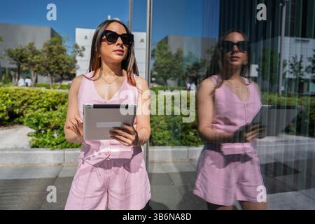 Junge Geschäftsfrau, die ein digitales Tablet in der Stadt verwendet, ihr Bild spiegelt sich in einem Glasfenster wider Stockfoto