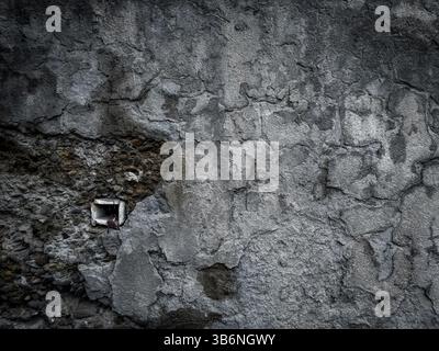 Beschädigte Zementwand mit freiliegendem Loch und robuster Textur, mit verwittertem Mauerwerk, abgebrochenem Putz und sichtbaren Körnerdetails Stockfoto
