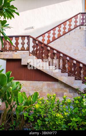 Elegante Steintreppe mit kunstvollen Holzbalustraden im balinesischen Stil, umgeben von üppigen tropischen Pflanzen und weißen Wänden. Stockfoto