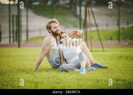 Sport und Technik. Junge in der Liebe heterosexuellen Kaukasischen paar Ausruhen nach Training draußen im Park auf Rasen, grünes Gras sitzen in Umarmung und m Stockfoto