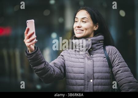 Nahaufnahme einer lächelnden, schönen Geschäftsfrau mittleren Alters, die selfie mit verschwommener Sicht im Hintergrund auf das Smartphone nimmt Stockfoto