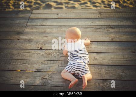 Neugeborene Kind ein Junge, ein Jahr alt, liegt auf seinem Bauch auf ein hölzernes Dock, einem Pier in gestreifter Kleidung, eine Sicht von oben im Sommer eine komplet bei Sonnenuntergang Stockfoto