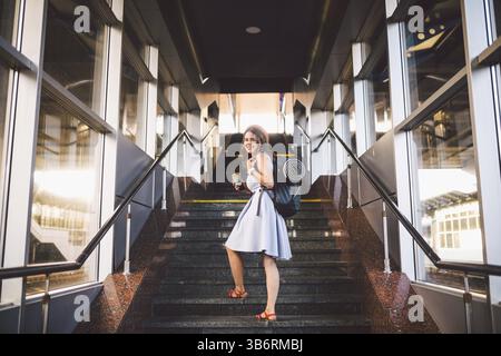 Junge schöne kaukasische Touristin in Sommerkleid und Hut mit Touristenrucksack klettert die Sommerfrau am Bahnhof hinauf. Schüler g Stockfoto