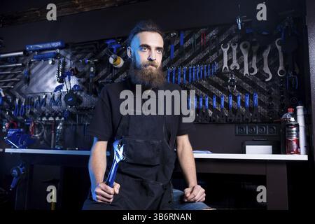 Portrait von Small Business Owner junger Mann mit Bart. Guy Fahrradmechaniker workshop Arbeiter sitzen mit Werkzeug in der Hand in einem schwarzen Kleide Stockfoto