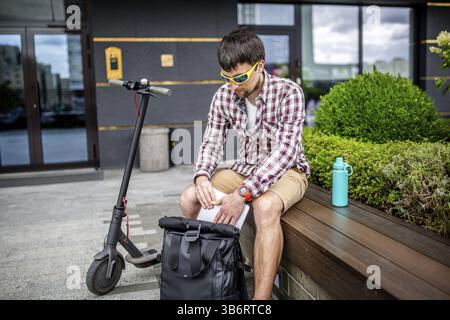 Gutaussehender Bärtiger im Hipster-Stil arbeitet online Freelancer, zieht Laptop mit Rucksack und Elektroroller aus, aktiver Lebensstil, Rucksackreisen Stockfoto