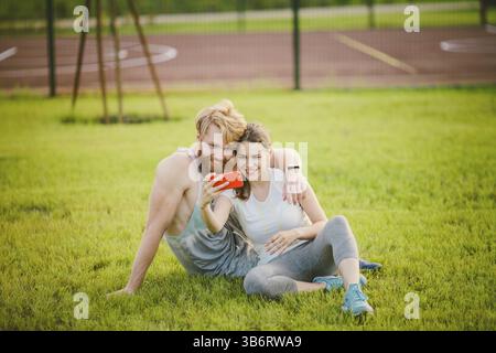 Sport und Technik. Junge in der Liebe heterosexuellen Kaukasischen paar Ausruhen nach Training draußen im Park auf Rasen, grünes Gras sitzen in Umarmung und m Stockfoto
