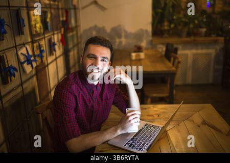 Eine junge Stattlichen kaukasischen Mann mit Bart und toothy Lächeln in rot kariertem Hemd arbeitet hinter Laptop an den hölzernen Tisch sitzen. Verwendet hält Mobile Stockfoto