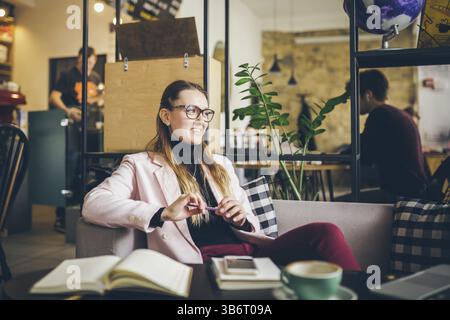 Träumende hübsche Dame, die an Ideen denkt, die im Café sitzen, kreativer Beruf. Thema moderne Bloggerin. Junge Frau mit Stift in der Hand, die an PLA denkt Stockfoto
