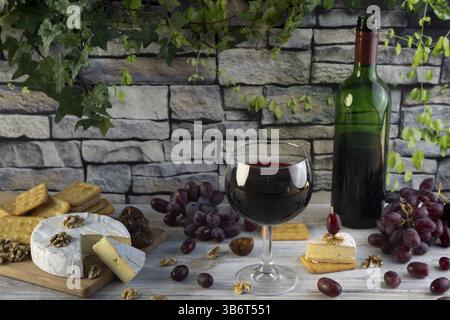 Weinglas und Flasche Rotwein umgeben von Käse, Trauben, Nüssen und Crackern vor einer rustikalen Steinmauer Stockfoto