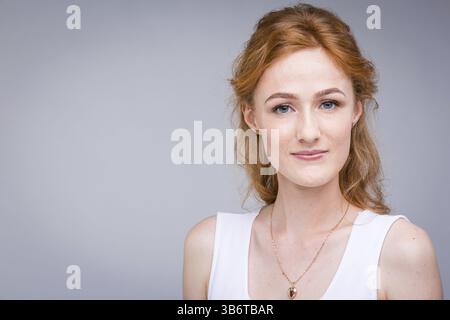 Nahaufnahme Porträt junge, schöne Geschäftsfrau, Studentin mit gelockten, lockigen Haaren und Sommersprossen auf Gesicht auf grauem Hintergrund im Studio. In weiß gekleidet Stockfoto