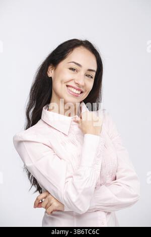 Porträt einer jungen Frau mit einem charmanten zahnigen Lächeln, schwarzen Haaren und braunen Augen auf weißem Hintergrund in einem rosa Hemd. Positive und freudige Emotionen Stockfoto