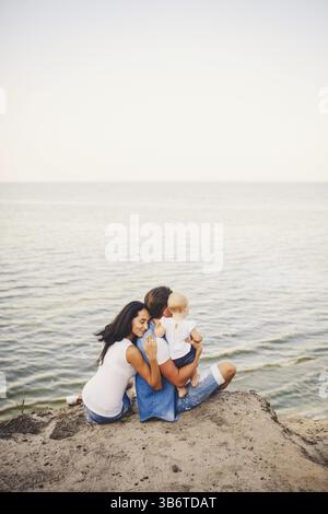 Themenurlaub mit Kleinkind in Natur und Meer. Mom, Dad und Tochter von einem Jahr sitzen in Umarmung, Mädchen in den Händen eines Mannes, mit Stockfoto