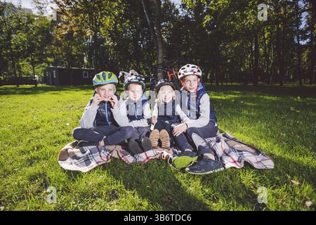 Große Familie kaukasische Kinder. Drei Brüder und Schwester sitzen auf einer Decke vor dem Park. Grüner Rasen Gras Hintergrund von Fahrrädern. Lustig Stockfoto