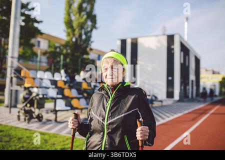 Aktive ältere kaukasische ältere Frauen von 90 Jahren üben Nordic Walking mit Skistöcken auf einer Strecke mit roter Gummibeschichtung. Aktivurlaub. Eine Passform Stockfoto