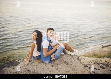 Themenurlaub mit Kleinkind in Natur und Meer. Mom, Dad und Tochter von einem Jahr sitzen in Umarmung, Mädchen in den Händen eines Mannes, mit Stockfoto