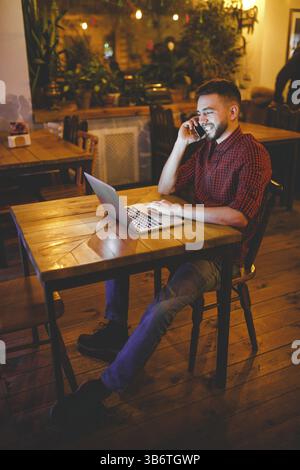 Junge stattlichen kaukasischen Mann mit Bart und toothy Lächeln im roten Hemd arbeitet hinter Laptop, Hände auf der Tastatur an den hölzernen Tisch sitzen. Verwendet, um die Anrufe auf Mo Stockfoto
