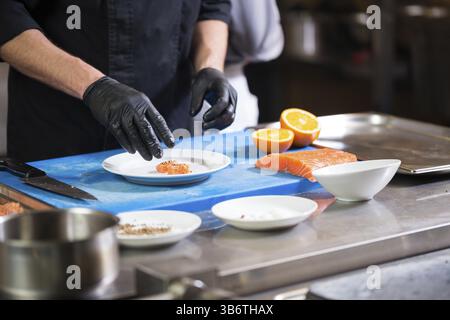 Thema Kochen ist ein Beruf des Kochens. Nahaufnahme der Hand ein kaukasischer Mann in einem Restaurant Küche vorbereiten rot Fischfilets Lachs Fleisch in schwarz l Stockfoto