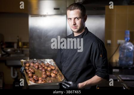 Portrait männlicher Koch mit gekochten Speisen in der Küche. Themenkochen. Junger kaukasischer Mann in schwarzer Uniform, Latexhandschuhe im Restaurant in der Küche. d Stockfoto