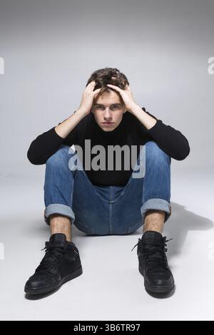 Ein stylischer Kerl posiert im Studio und sitzt auf dem Boden in Jeanshosen und schwarzem Sweatshirt. Männliches Studentenporträt voll auf weißem Hintergrund. Studio Shot Han Stockfoto