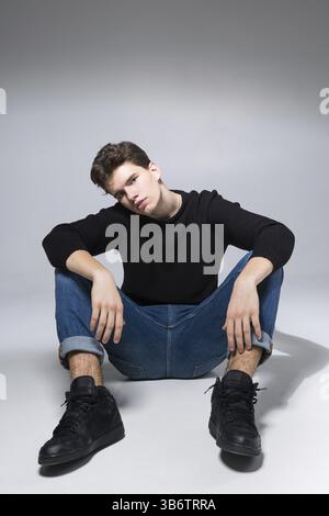 Stilvoller, gutaussehender Mann, der auf dem Boden sitzt. Gutaussehender Guy. Cooles modisches männliches Modell, das auf grauem Hintergrund sitzt und die Kamera betrachtet. Student in A. Stockfoto