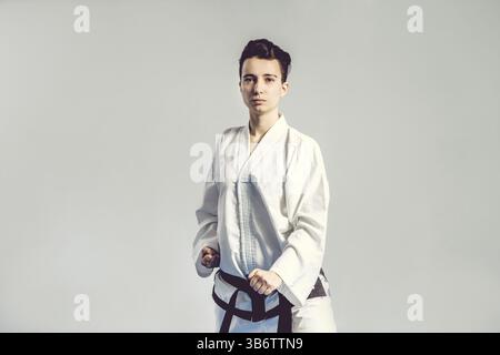 Mädchen im Karate-Anzug-Kimono im Studio auf grauem Hintergrund. Das weibliche Kind zeigt Judo oder Karate stans in weißer Uniform mit schwarzem Gürtel. Individual Martial Stockfoto