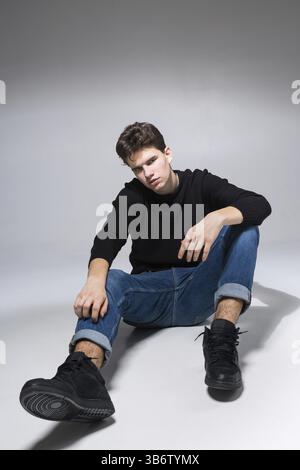 Ein stylischer Kerl posiert im Studio und sitzt auf dem Boden in Jeanshosen und schwarzem Sweatshirt. Männliches Studentenporträt voll auf weißem Hintergrund. Studio Shot Han Stockfoto