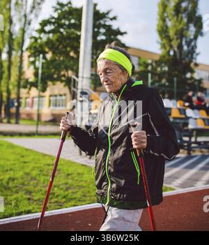 Ältere Frau, die mit Gehstöcken im Stadion auf einer roten Gummihülle läuft. Ältere Frau 88 Jahre alt, die Nordic Walking Übungen im stadiu macht Stockfoto