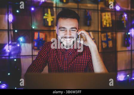 Junge stattlichen kaukasischen Mann mit Bart und toothy Lächeln im roten Hemd arbeitet hinter Laptop, Hände auf der Tastatur an den hölzernen Tisch sitzen. Verwendet, um die Anrufe auf Mo Stockfoto