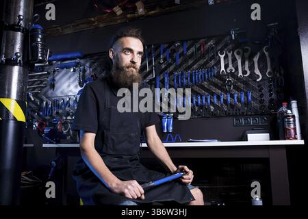 Portrait von Small Business Owner junger Mann mit Bart. Guy Fahrradmechaniker workshop Arbeiter sitzen mit Werkzeug in der Hand in einem schwarzen Kleide Stockfoto