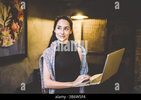 Das Konzept des Kleinunternehmens und der Technologie. Junge schöne Brünette Geschäftsfrau in schwarzem Kleid und grauem Pullover steht im Café mit Laptop Stockfoto