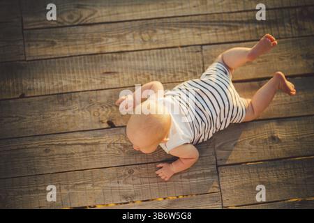 Neugeborene Kind ein Junge, ein Jahr alt, liegt auf seinem Bauch auf ein hölzernes Dock, einem Pier in gestreifter Kleidung, eine Sicht von oben im Sommer eine komplet bei Sonnenuntergang Stockfoto