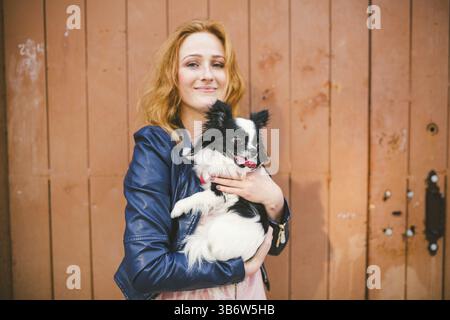 Junge kaukasische rothaarige Frau, die einen Hund der Rasse Chihuahua auf hölzernem Hintergrund an den Händen hält. Das Thema Tierliebe, Schutz des pe Stockfoto