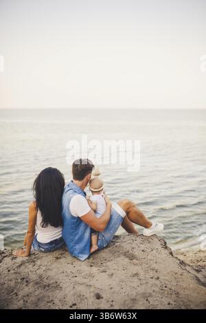 Themenurlaub mit Kleinkind in Natur und Meer. Mom, Dad und Tochter von einem Jahr sitzen in Umarmung, Mädchen in den Händen eines Mannes, mit Stockfoto