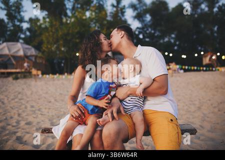 Das Konzept eines Familienurlaubs. Junge Familie und zwei Söhne sitzen abends auf einer Bank am Sandstrand. Mama und Dad küssen sich, der ältere Bruder kis Stockfoto