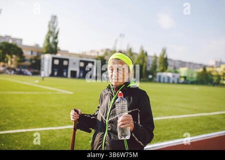 Eine alte Frau, die mit nordic Walk Stöcken auf der Laufbahn lief, ein Gummiband und hielt an, um Durst zu stillen, Wasser aus einer Flasche zu trinken. Aktives weibliches SEN Stockfoto