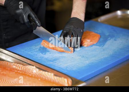 Thema Kochen ist ein Beruf des Kochens. Nahaufnahme der Hand ein kaukasischer Mann in einem Restaurant Küche vorbereiten rot Fischfilets Lachs Fleisch in schwarz l Stockfoto