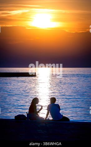 Eine romantische Sonnenuntergangsszene mit zwei Silhouetten, die am Wasser sitzen und die Gesellschaft des anderen genießen. Die Sonne untergeht am Horizont und wirft einen warmen Glo Stockfoto