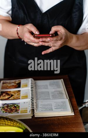 Frau zu Hause in der Küche mit schwarzer Schürze, die ein Foto von einem Rezept aus einem Rezeptbuch macht Stockfoto