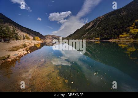 Kristallklarer Bergsee, der den dramatischen Himmel reflektiert, und immergrüne Hänge, umgeben von einer ruhigen Herbstkulisse. Stockfoto