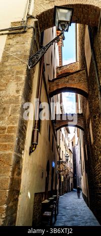 Eine enge Straße in einer Altstadt mit Steinhäusern und Bögen. Das Sonnenlicht filtert durch die Bögen und schafft eine warme Atmosphäre. Eine Person Stockfoto